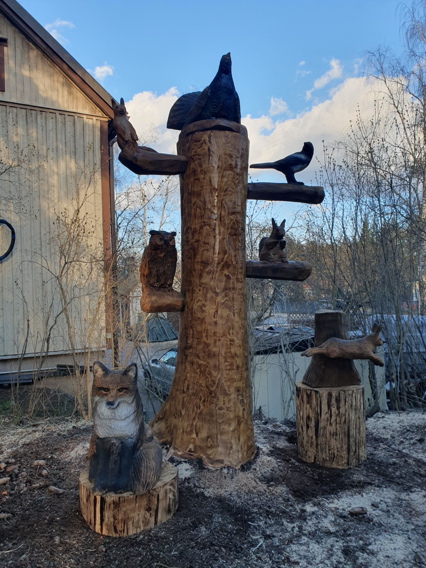 Pihapiirissä puuveistoksia. Veistoksessa on kettu, loikkaava jänis, kaksi oravaa, pöllö, harakka ja veistoksen päällä metso. Taustalla sininen taivas ja pensaita.
