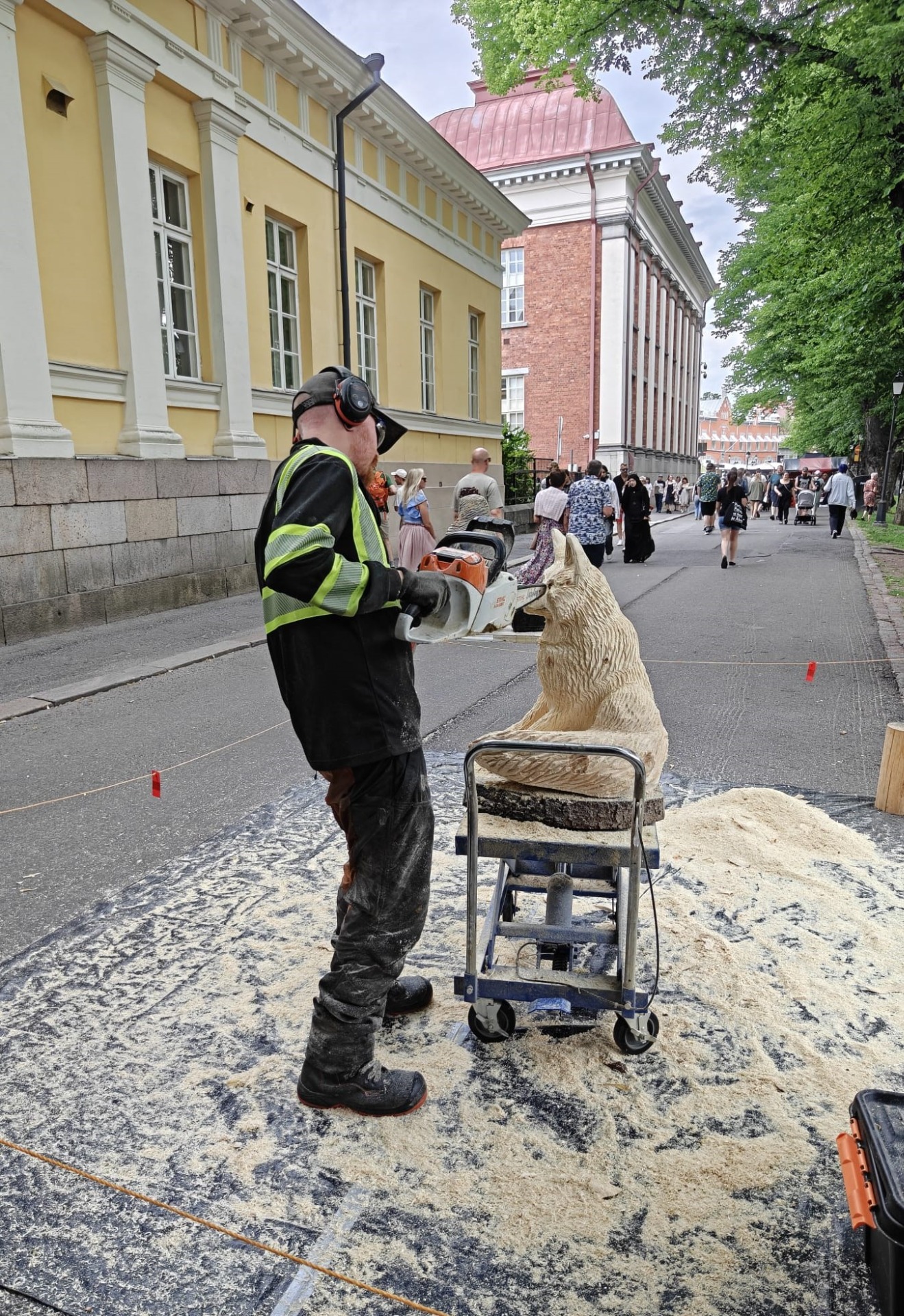 Katuympäristössä puuveistäjä veistää veistosta. Taustalla yleisöä ja historiallisia rakennuksia.
